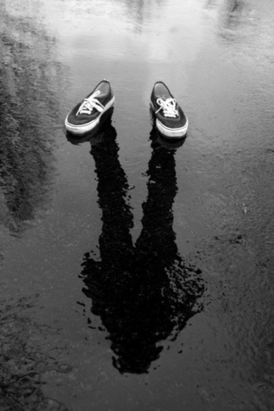 A worn pair of shoes beside the reflection of a person in a puddle.